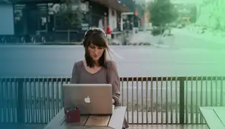 Bild zeigt eine Frau, die mit dem Laptop an einem Tisch draussen auf einer Bank arbeitet.