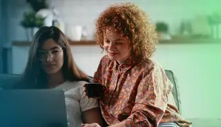 Header zeigt zwei Mitarbeiterinnen mit Laptop und Kaffeebecher, die Pause machen.