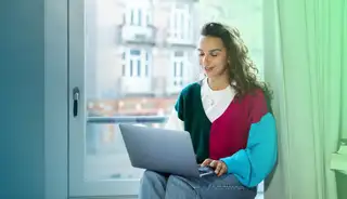 Frau mit Laptop sitzt auf Fensterbank und arbeitet mit askDANTE zur Gleitzeit.