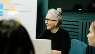 Headerbild zeigt eine lachende Frau, die mit ihrem Laptop in einem Meeting sitzt und ihren Stundennachweis automatisch mit askDANTE erbringt.