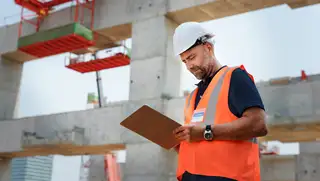 Headerbild zeigt einen Mitarbeiter in Zeitarbeit auf der Baustelle, der seine Arbeitszeit mit askDANTE erfasst hat und seine Aufgaben am Klemmbrett durchgeht.