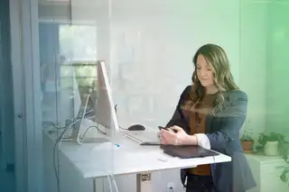 Headerbild zeigt Mitarbeitern am Schreibtisch, die vor einem Monitor steht und am Handy einen Urlaubsantrag stellt.