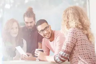 Bild zeigt vier Personen, die sich mit einer Kaffeetasse, einem Tablet und einem Stift über einen Tisch lehnen.