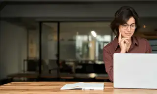 bild zeigt einen schreibtisch aus holz von vorne an dessen rechter seite eine frau mit brille am laptop sitzt